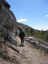 Hiker walks along dirt trail along rocky cliff on one side and rocks into green hills on the other.