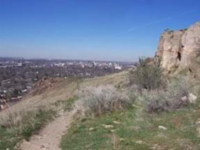 View of Boise from Table Rock with dirt trail, grass and cliff in image.